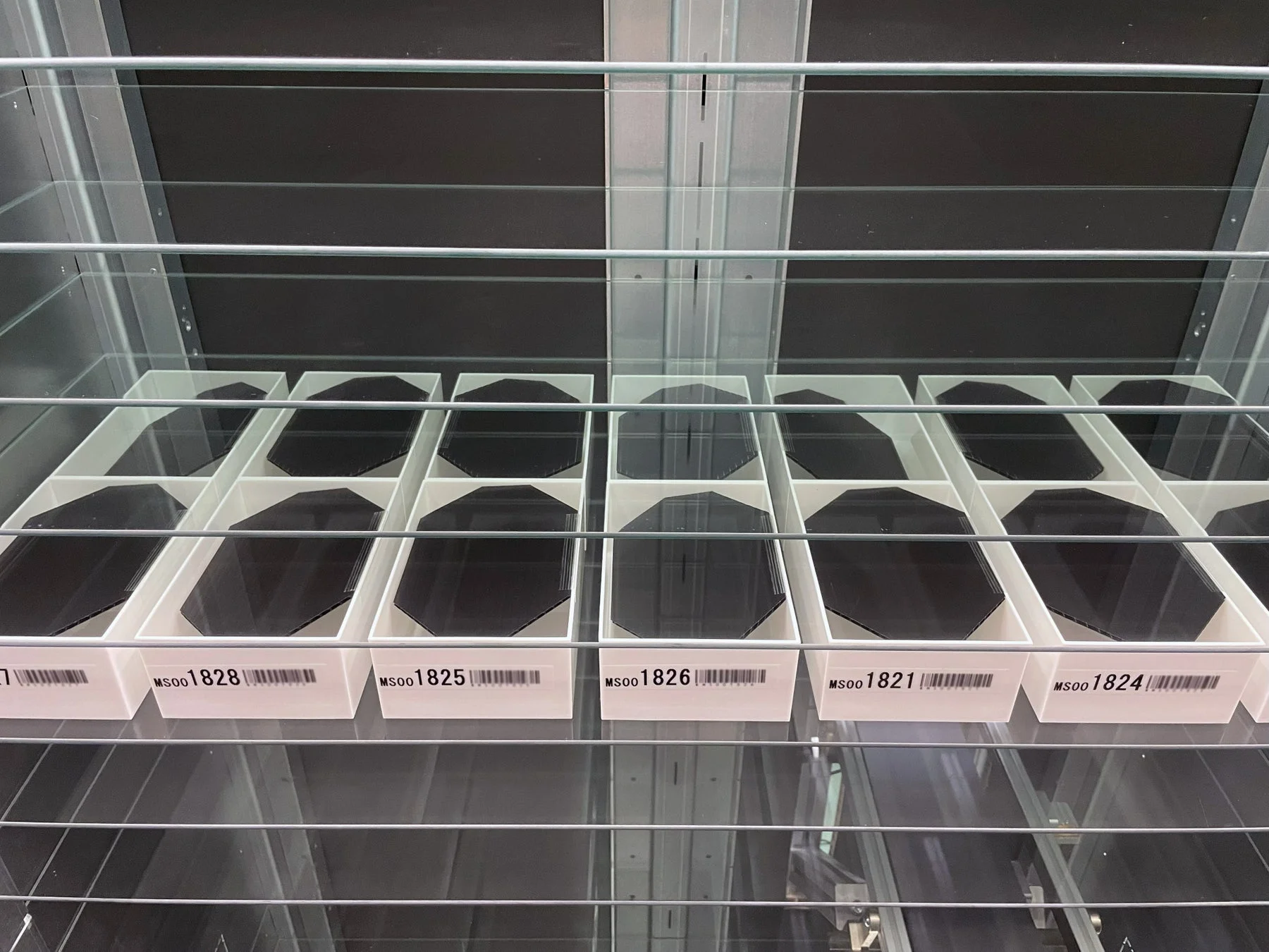 ガラス棚に並べられた白いトレイの中に、黒い多角形の物体が整然と保管されている様子を捉えた画像です。各トレイには識別用のバーコードと番号が記載されています。半導体ウェハーのような工業用または研究用のサンプル保管庫と推測されます。