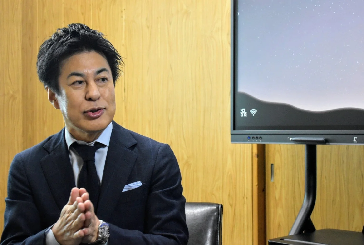 ダークスーツを着たアジア人男性が、木目調の壁と大型モニターの前で手を組みながら話している。ビジネスシーンでの会議やプレゼンテーションを思わせる一枚。