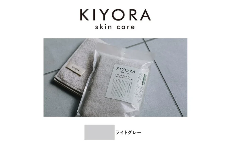 KIYORA skin careのライトグレーのタオルが写っています。折りたたまれたタオルと、パッケージに入った「今治生まれの洗顔専科 こすらないスキンケアタオル」が床に置かれており、清潔感のある印象です。