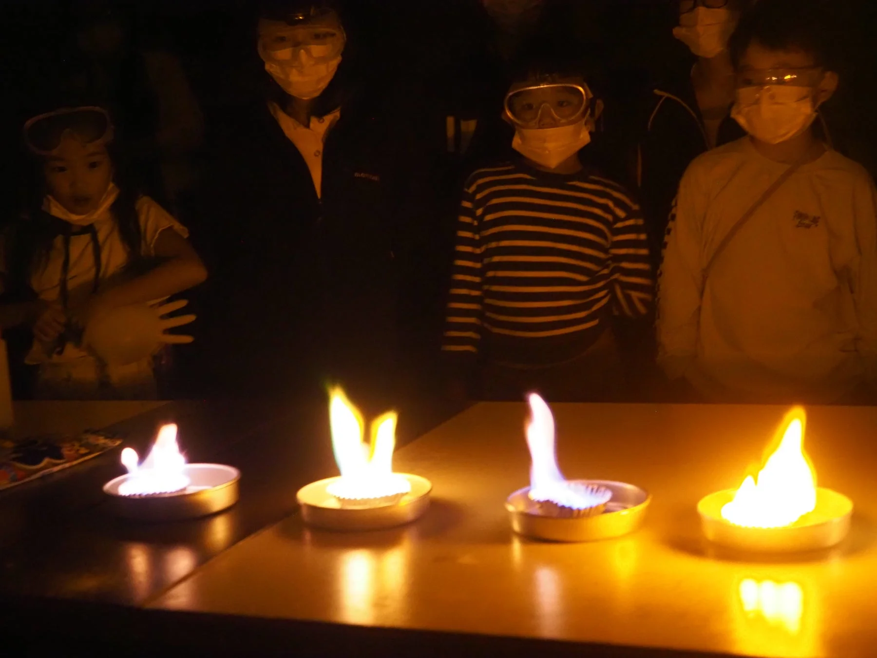 暗い部屋で、安全ゴーグルとマスクを着用した子供たちが、炎色反応の科学実験を観察しています。テーブルの上には複数の異なる色の炎が燃えています。