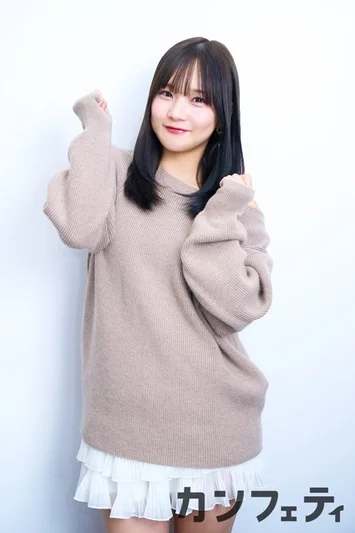 白い背景のスタジオで、笑顔でポーズをとる若い日本人女性のポートレート。ベージュのオーバーサイズニットと白いフリルスカートを着用しています。