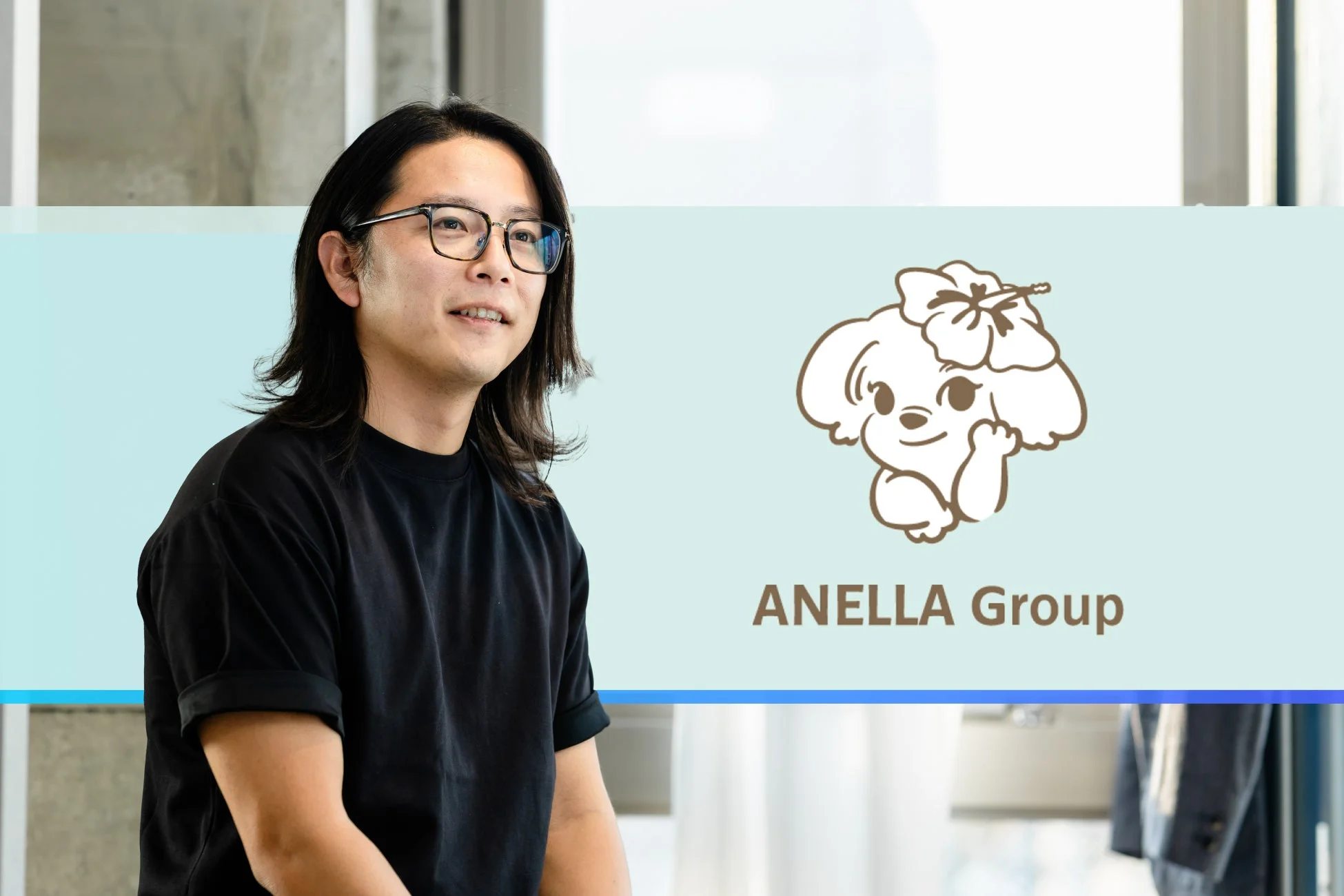 長髪とメガネの男性が黒いTシャツ姿で写っており、背景には犬のキャラクターと「ANELLA Group」という企業ロゴが配置されています。
