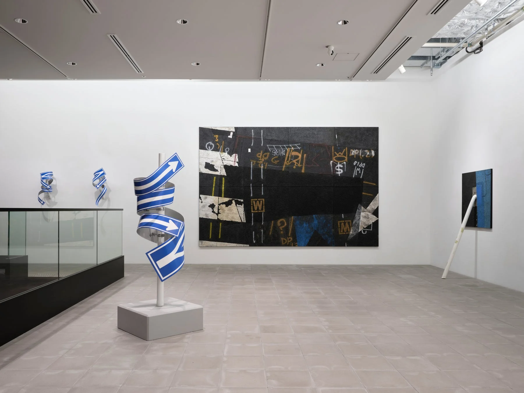 現代アートギャラリーの展示風景。ひねられた道路標識のような青い矢印の彫刻と抽象画。壁にはグラフィティのような文字や記号が描かれ、全体的にミニマルな空間に複数の現代美術作品が展示されています。