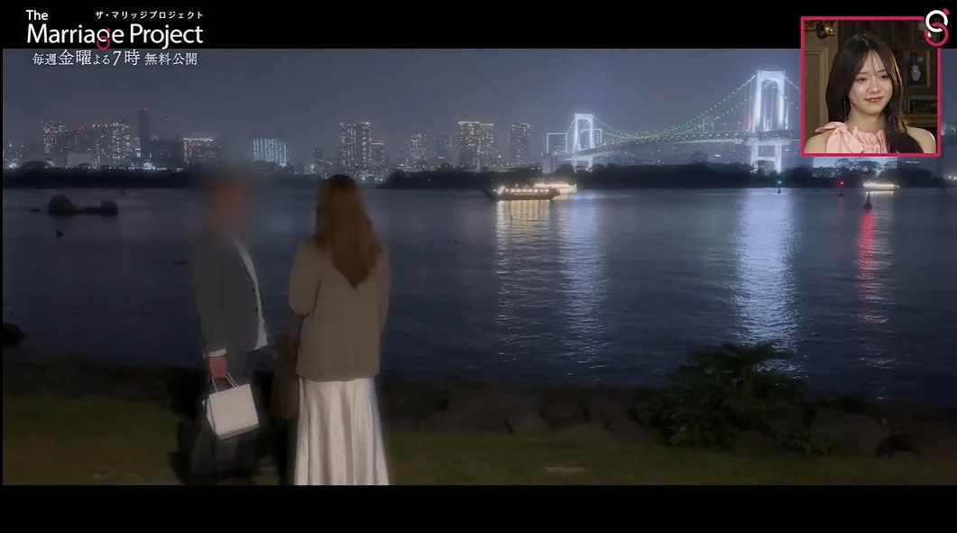 夜景を背景に、水辺で男女が向かい合って立っている。遠くには煌めく都市のビル群とライトアップされた橋が見える。画面右上には女性の顔が映る小窓がある。