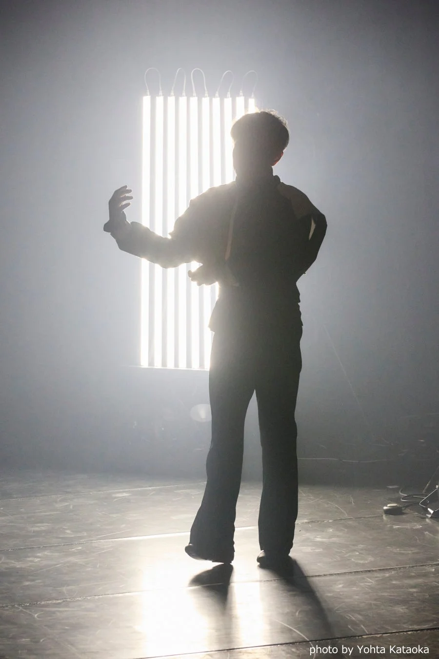 霧のかかったステージで、明るい縦型ライトを背に立つ人物のシルエット。幻想的な光の演出が際立つ、神秘的なパフォーマンスの一場面を捉えた写真です。