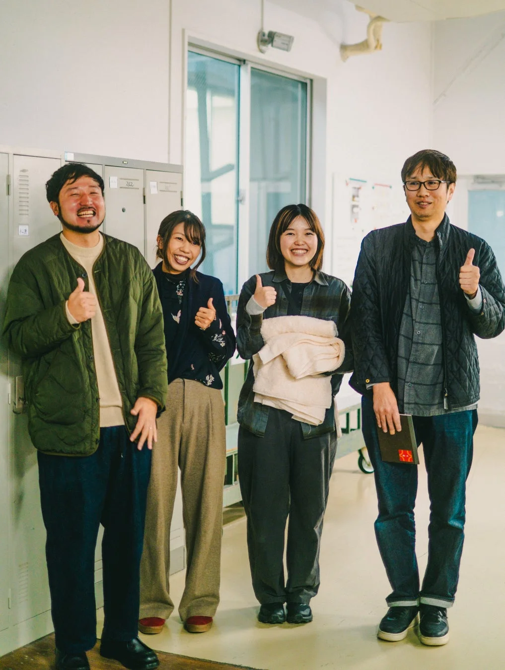 屋内で4人の人々が笑顔でサムズアップしています。オフィスまたは学校のような場所で、皆が明るく前向きな雰囲気です。