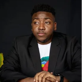 黒い背景の前で、黒いスーツジャケットとカラフルな模様の白いTシャツを着た男性が、少し横を向いて座っているポートレートです。真剣な表情をしています。