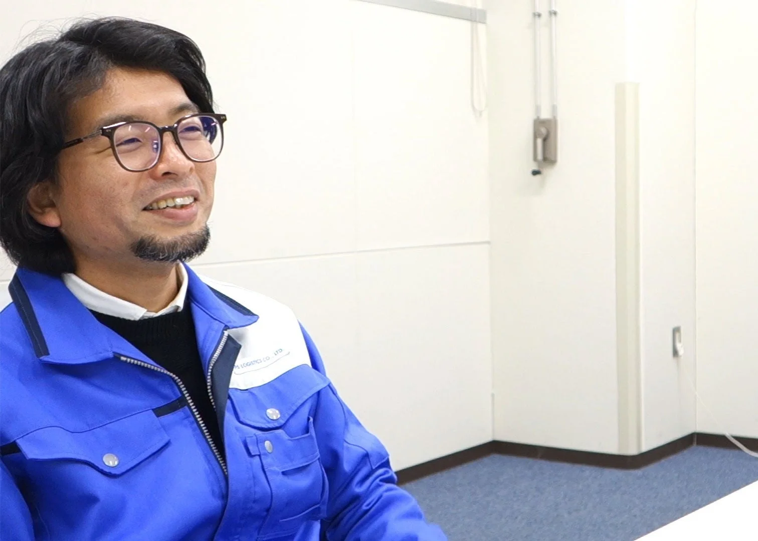 青い作業着を着用した男性が、室内で優しい表情で右方向を見ている様子を捉えた画像です。メガネとひげが特徴的です。