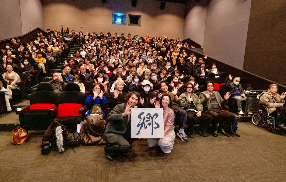 映画館で大勢の観客が手を振っており、前列の2人が「郷 THE HOMECOMING」と書かれたボードを掲げています