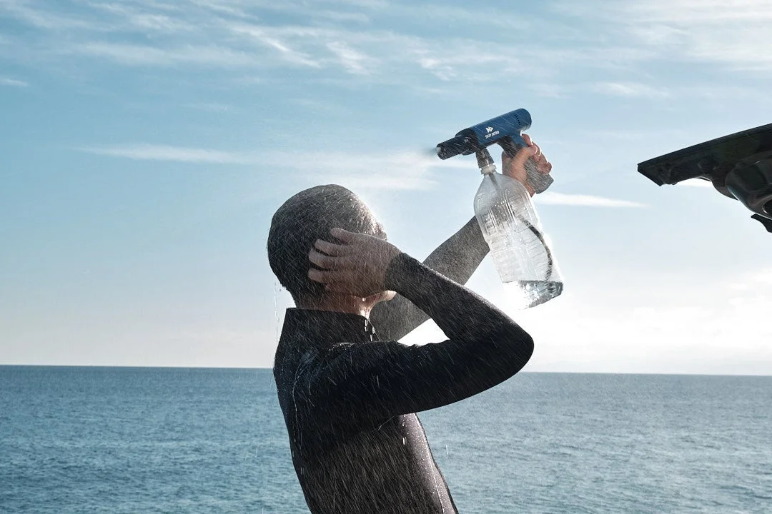 ウェットスーツを着た人が、海を背景にポータブルシャワーを使って体を洗い流している様子