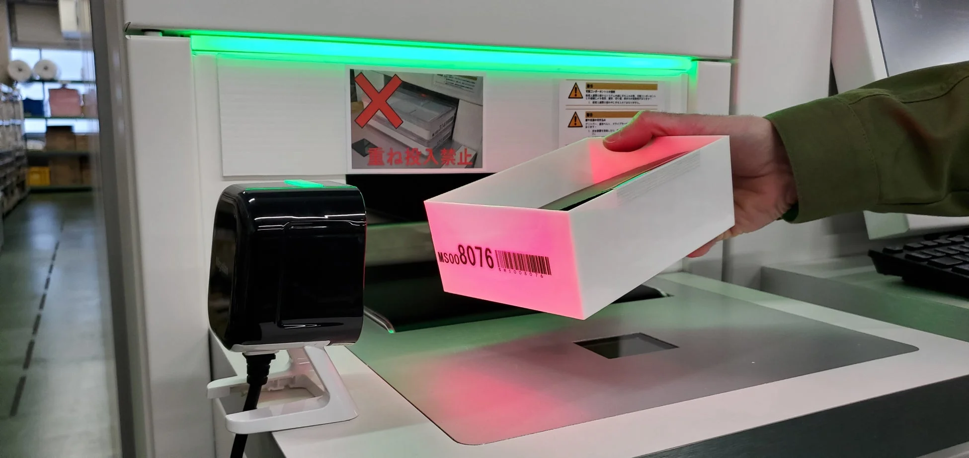 物流施設らしき場所で、作業員がバーコードリーダーを使って荷物（箱）をスキャンしている様子です。機械には「重ね投入禁止」の注意書きが見えます。