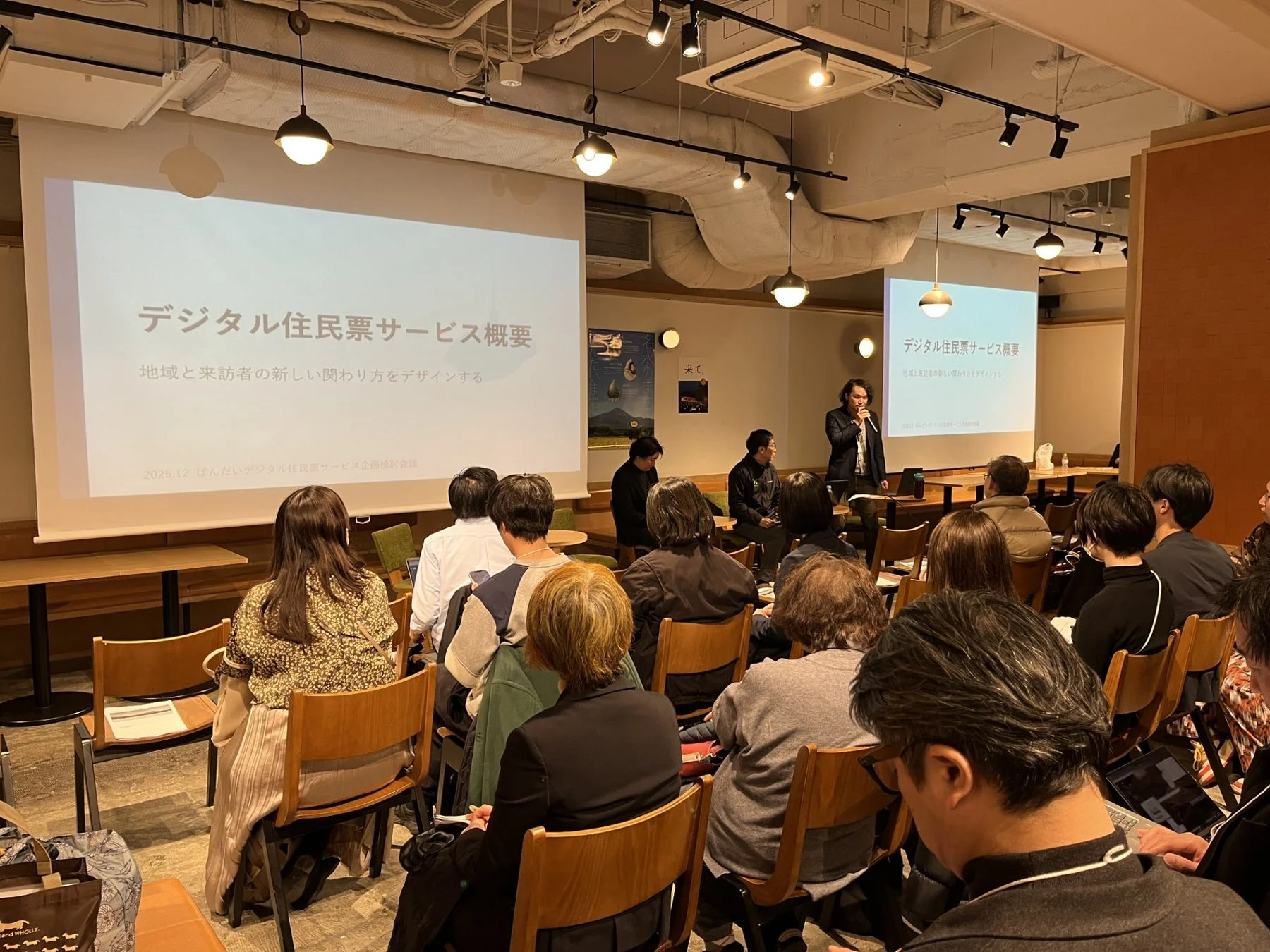 「デジタル住民票サービス概要」というテーマで講演が行われている会場の様子。登壇者がマイクを手に話し、多数の参加者が熱心に耳を傾けている。