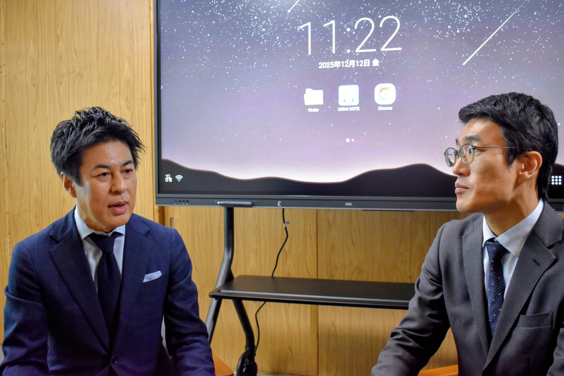 スーツを着た二人の男性が会議室のような場所で話しています。背景には時刻や日付、アプリケーションが表示された大型ディスプレイがあります。