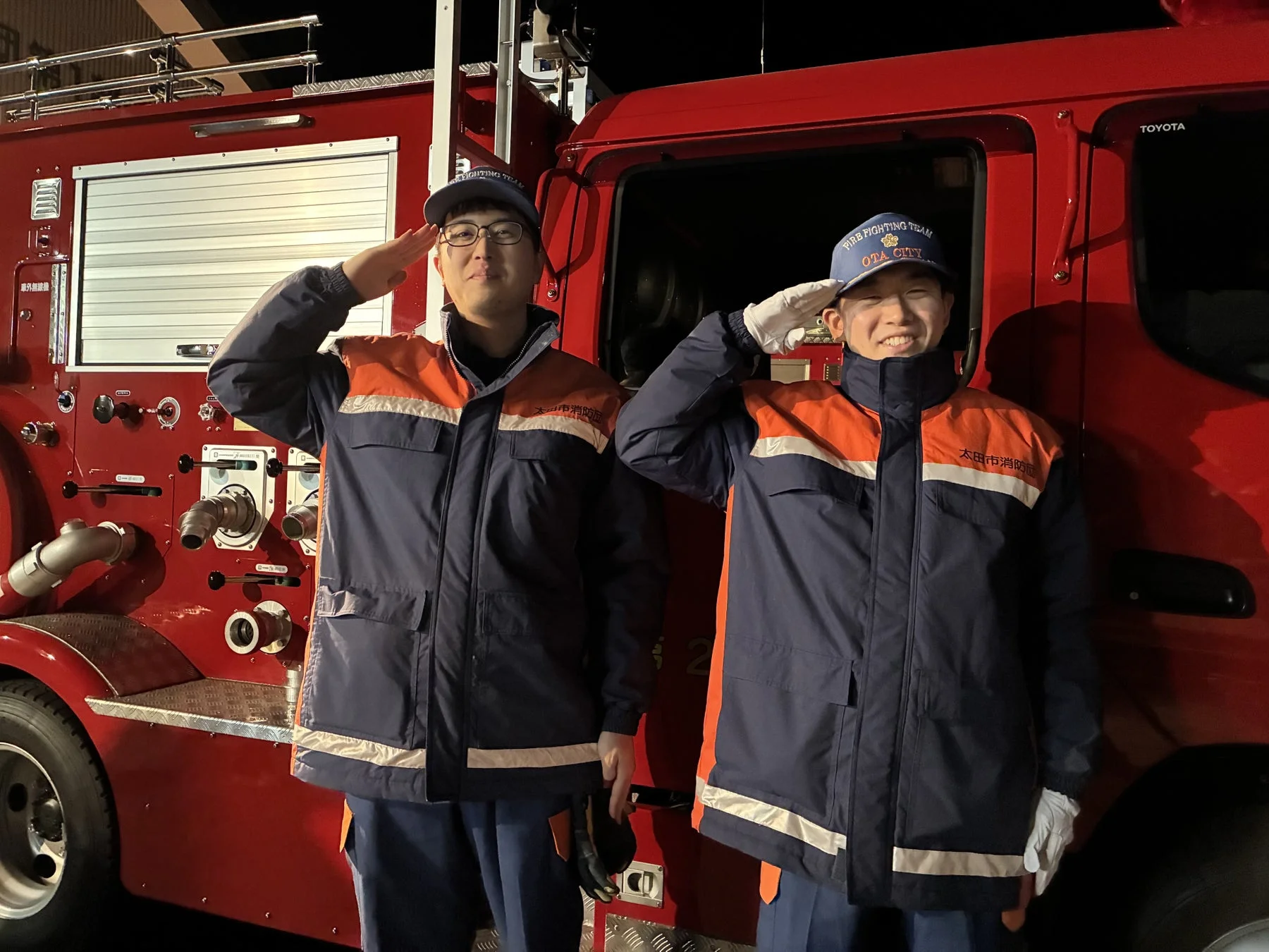 太田市消防の制服を着た2人の男性が、消防車の前で敬礼している夜間の写真。彼らは消防隊員で、地域社会の安全を守る役割を担っています。