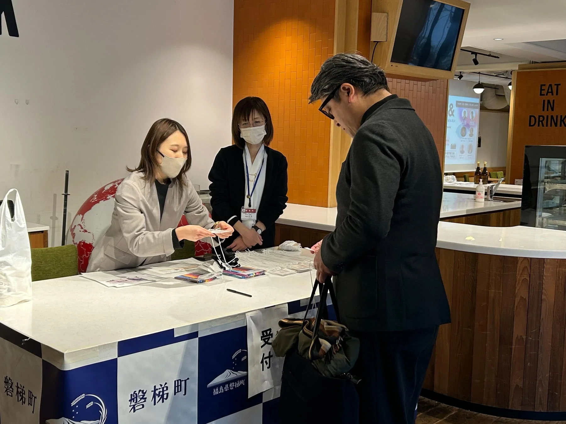 マスクを着用した2人の女性が受付ブースで男性に対応している様子。福島県磐梯町のPRイベントか物産展のような雰囲気の屋内会場である。