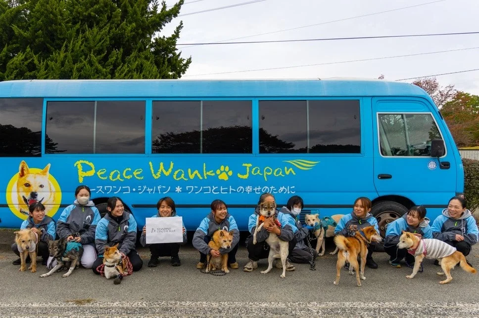 青いバスの前で集合写真に収まるピースワンコ・ジャパンのスタッフと犬たち