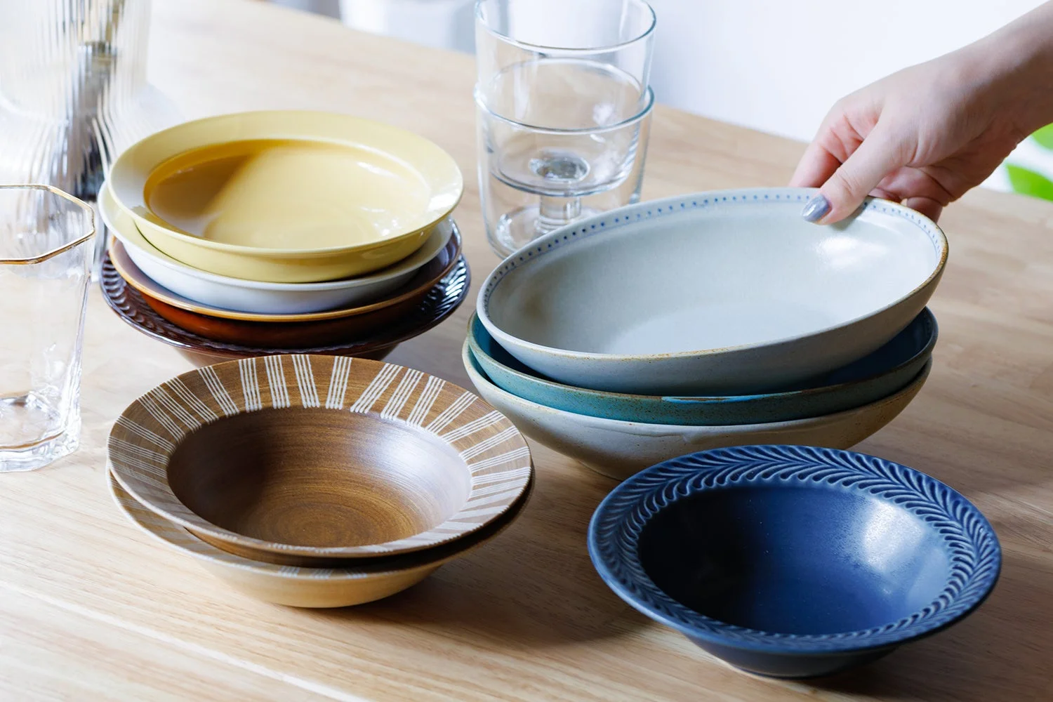 木製のテーブルに様々な色やデザインの陶器の皿、ボウル、グラスが並べられています。手で白いボウルを置いている様子が写っており、食卓を彩る食器のコレクションが魅力的な一枚です。