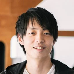 カメラに向かって微笑む若い日本人男性の顔のアップです。黒い髪で、明るい背景の前に立っています。