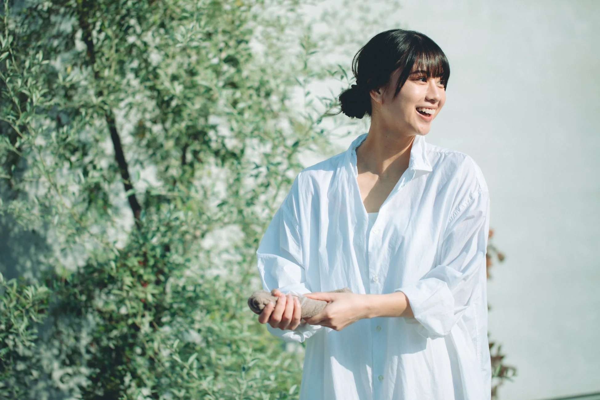 白いシャツを着た若い女性が、緑の木々と白い壁を背景に、明るい笑顔で横を向いています。自然光が差し込む屋外で、リラックスした爽やかな雰囲気が感じられるポートレートです。