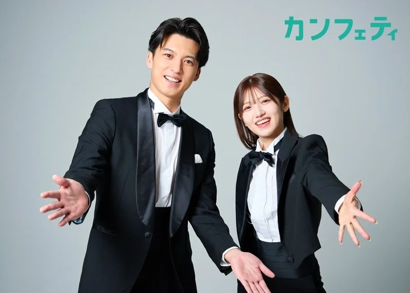 タキシード姿の男性と女性が、笑顔で両手を広げて観客を歓迎しているようなポーズをとっています。