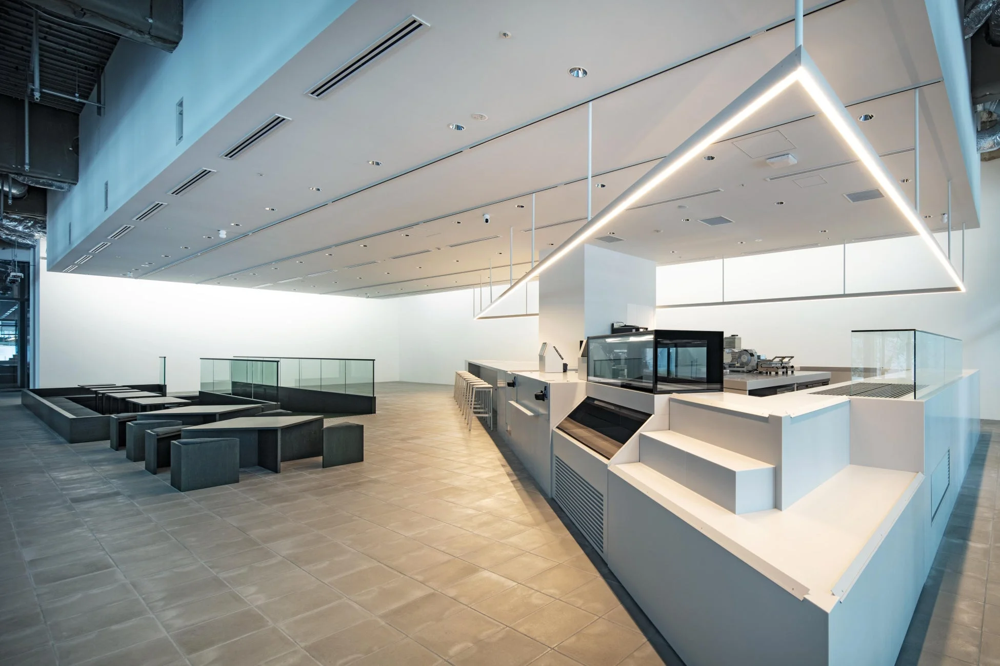 モダンでミニマリストなデザインのカフェまたはラウンジの室内空間。白いカウンター、黒い座席、そして天井からの特徴的な照明が配置され、広々とした印象を与える。