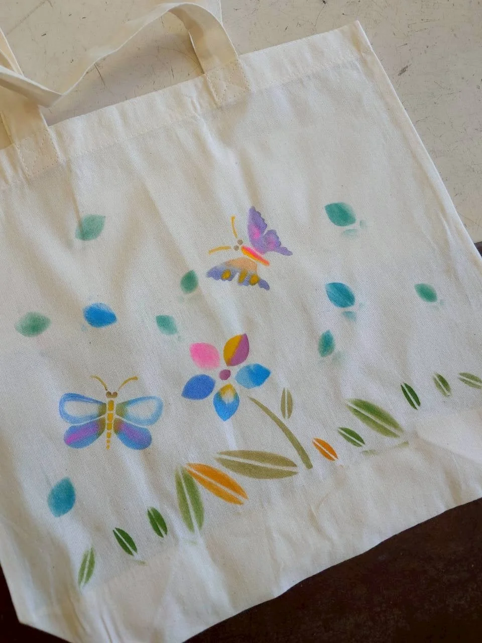 白地のトートバッグに、蝶や花、葉っぱのカラフルなステンシルアートが施されています。手作り感のある可愛らしいデザインです。