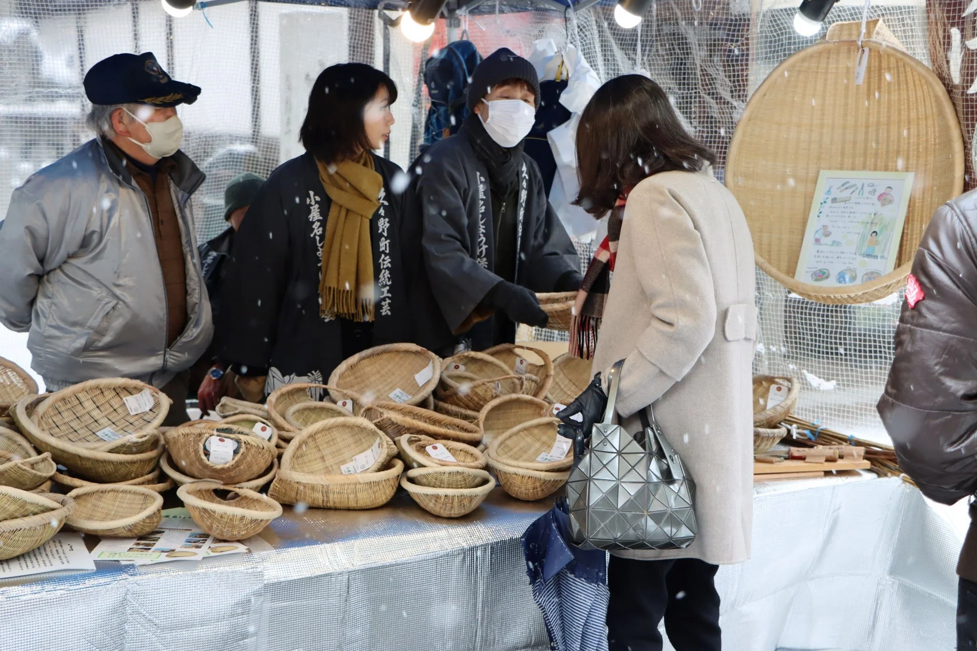 雪が舞う屋外の市場で、伝統的な手編みのかごを販売している様子。数人の人々が商品を見ていたり、店番をしていたりする。