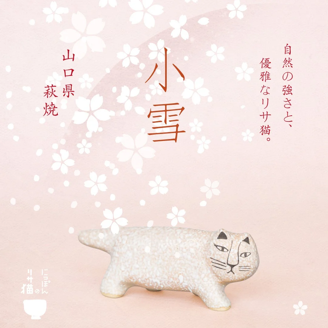 桜の花びらが舞う淡いピンクの背景に、白い猫の陶器の置物が描かれています。山口県の萩焼で、リサ・ラーソンの猫をモチーフにした「小雪」という作品のようです。自然の強さと優雅さを表現しています。