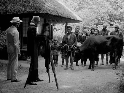 茅葺き屋根の家を背景に牛と人々が記念撮影をしている白黒写真