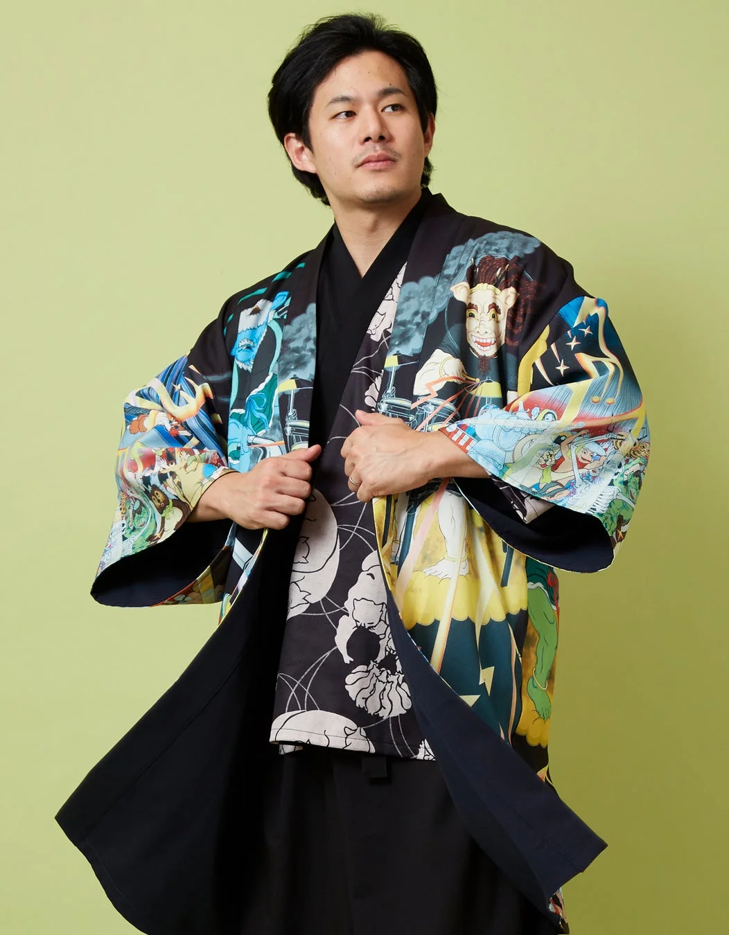 緑色の背景の前で、カラフルで個性的な柄の羽織を身につけた男性が、羽織を軽く開いてポーズをとっています。