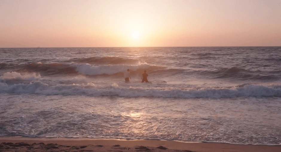 夕焼けに染まる海辺で、穏やかな波打ち際に立つ二人の姿