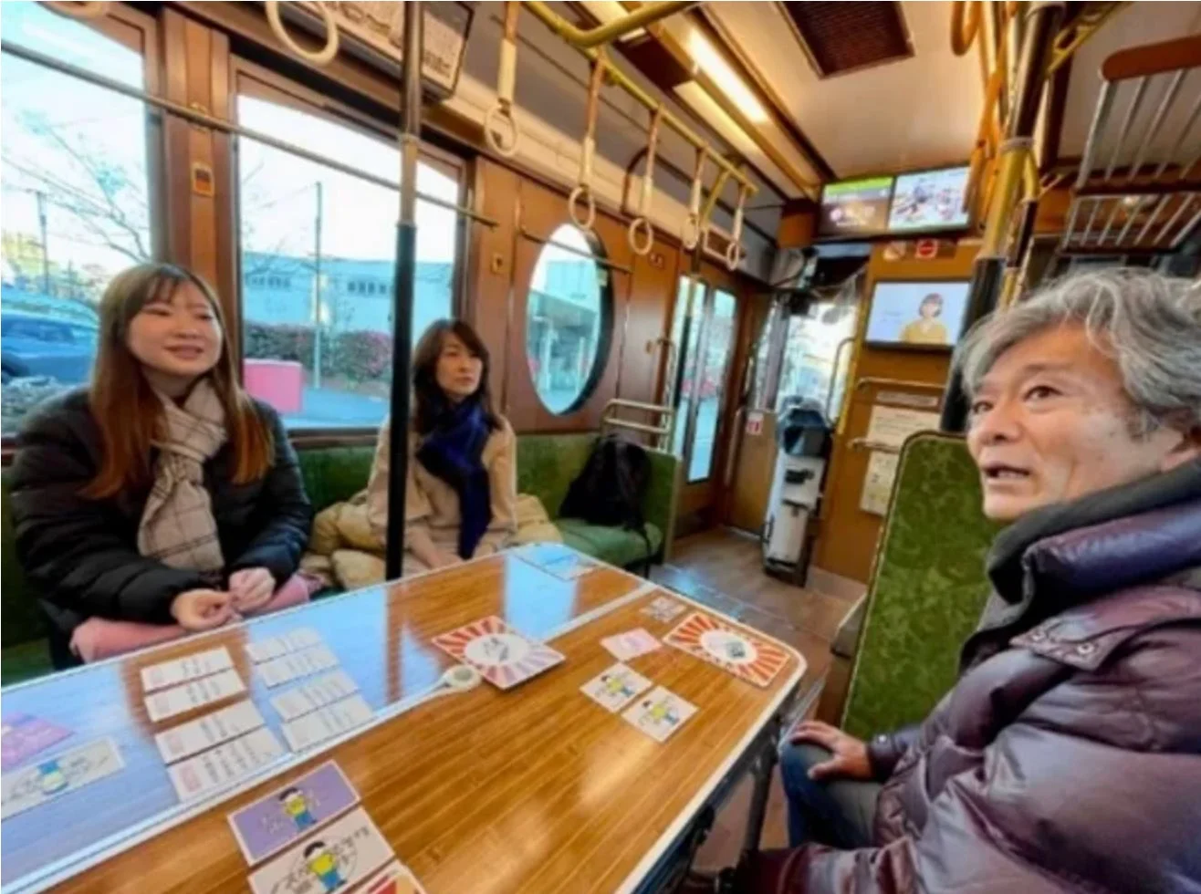 レトロな雰囲気の電車内で、3人の人々がテーブルを囲んでカードゲーム（かるた）を楽しんでいます。和やかな雰囲気で会話が弾んでいる様子がうかがえます。