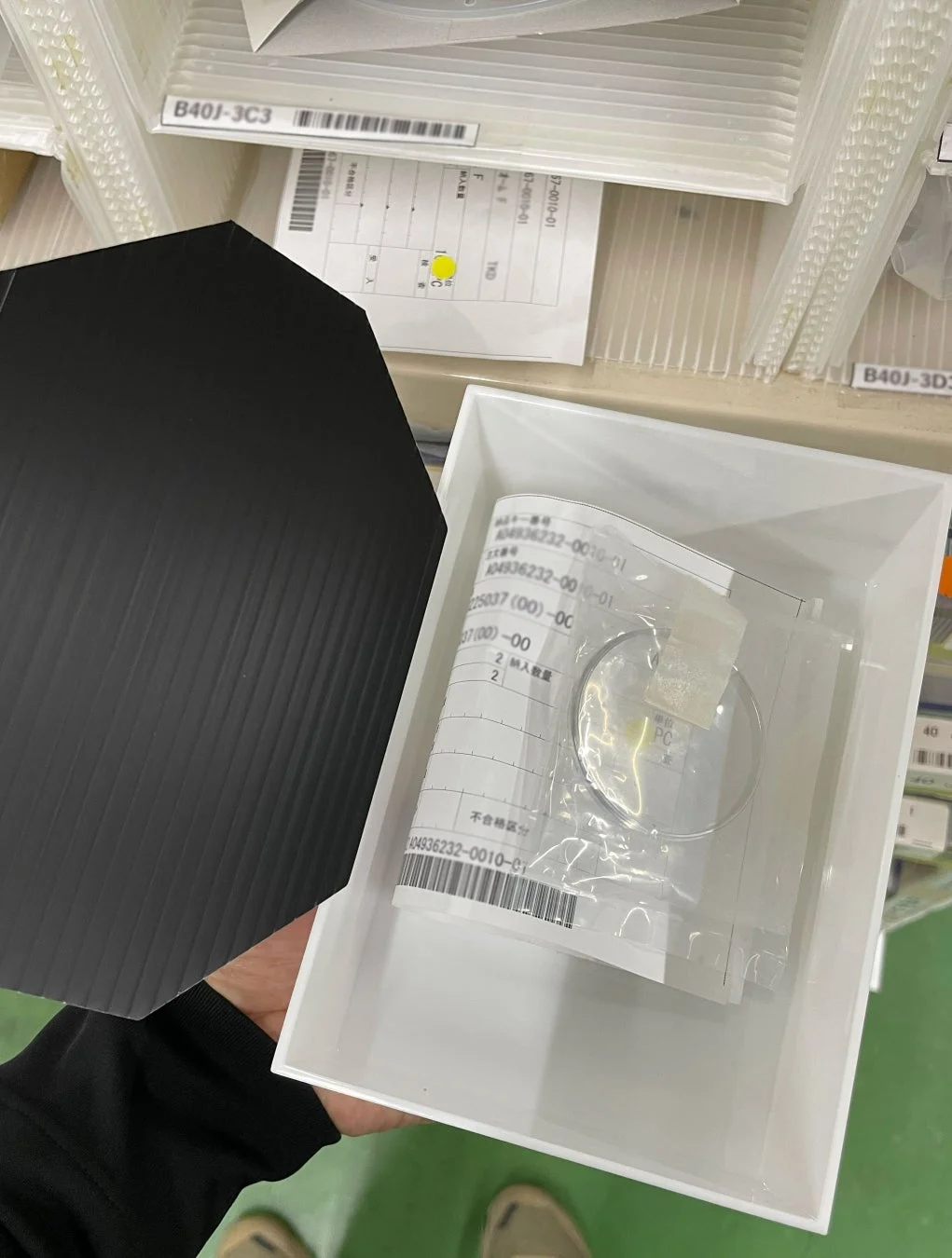 作業員が黒い六角形の部品と、書類や小さな金属リングが入った白いトレイを手にしています。背景には部品管理棚があり、製造現場や倉庫での部品取り扱い作業の様子がうかがえます。