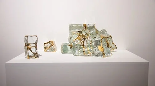 白い台座の上に置かれた、透明なガラスや樹脂に金箔が埋め込まれた現代アート作品のグループショット。