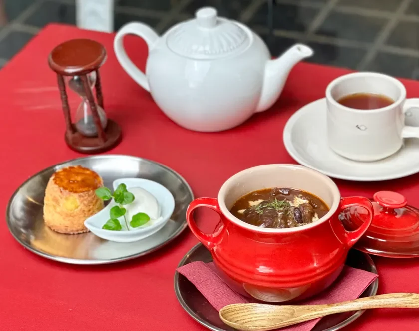 赤いテーブルクロスの上で、温かい紅茶とビーフシチュー、そしてスコーンが並べられたカフェでの食事風景です。砂時計も置かれており、ゆったりとした時間が流れている様子が伺えます。