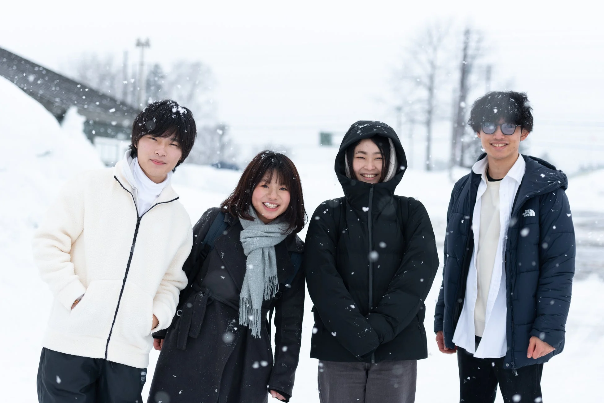 雪が降る中で、若者4人が笑顔で並んでいます。冬の屋外で楽しんでいる様子が伝わる集合写真です。