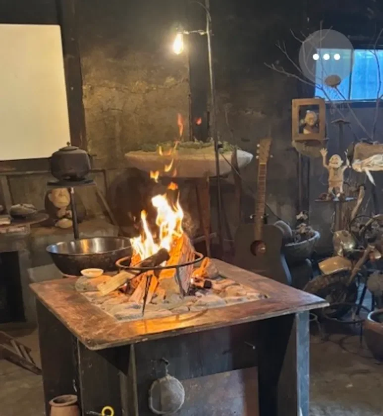 画像中央に焚き火台があり、燃え盛る炎が暖かさを感じさせます。周囲にはギター、古いケトル、猿の置物など、レトロで個性的な装飾品が配置されており、落ち着いた居心地の良い空間を演出しています。