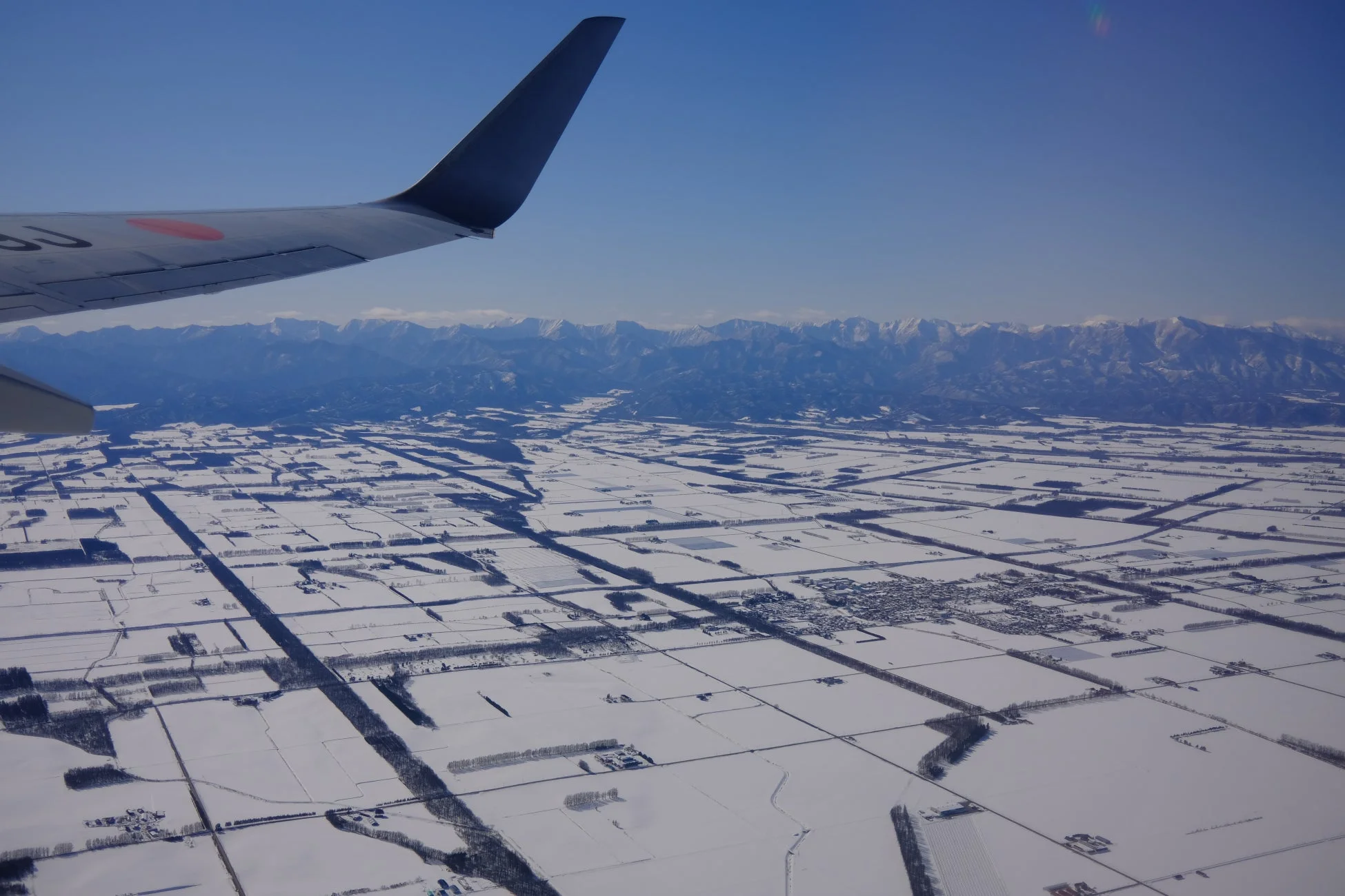 飛行機の窓から撮影された雪景色。手前に航空機の翼が見え、広大な雪に覆われた田園地帯が広がる。遠くには雪山が連なり、澄み切った青空が印象的な冬の空撮風景です。