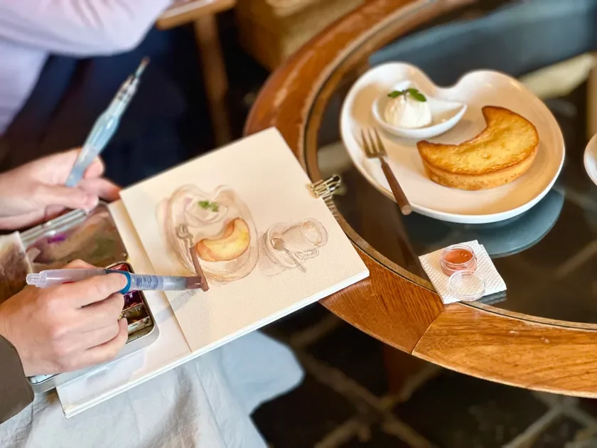 カフェで、目の前にある三日月型のケーキとアイスクリームのデザートを水彩画で描いている様子です。手元には絵の具パレットと水筆があり、絵が完成していく過程がうかがえます。