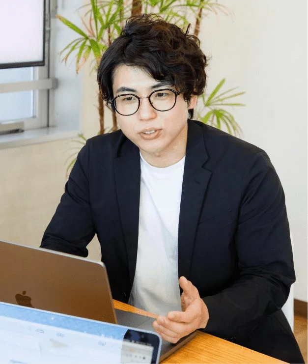 メガネをかけた若い男性が、オフィスらしき場所でノートパソコンを前に座り、何かを説明しているかのように話している様子