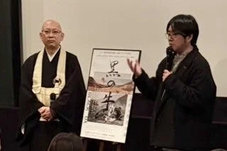 僧侶とスーツ姿の男性がマイクを持って話す様子