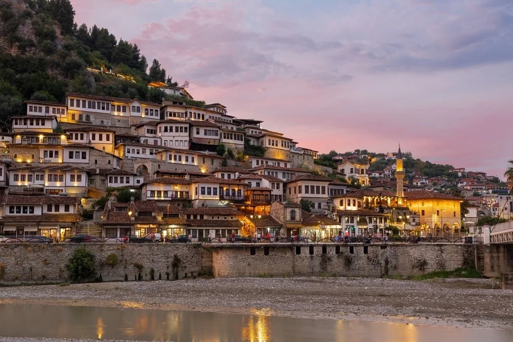 夕暮れ時、川沿いの急な斜面に白い壁と茶色の屋根の家々が密集して建ち並ぶ美しい街の風景