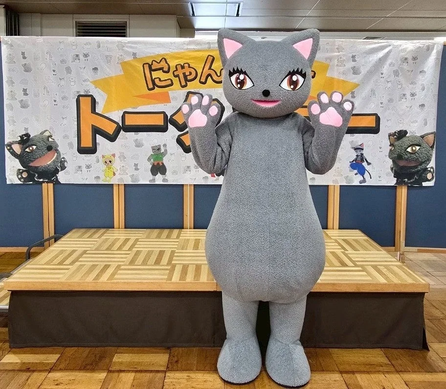 ステージ前に立つ灰色の猫の着ぐるみマスコットキャラクター。背景には「にゃん」という文字と猫のイラストが描かれたバナーがあり、イベント会場の雰囲気を伝えている。