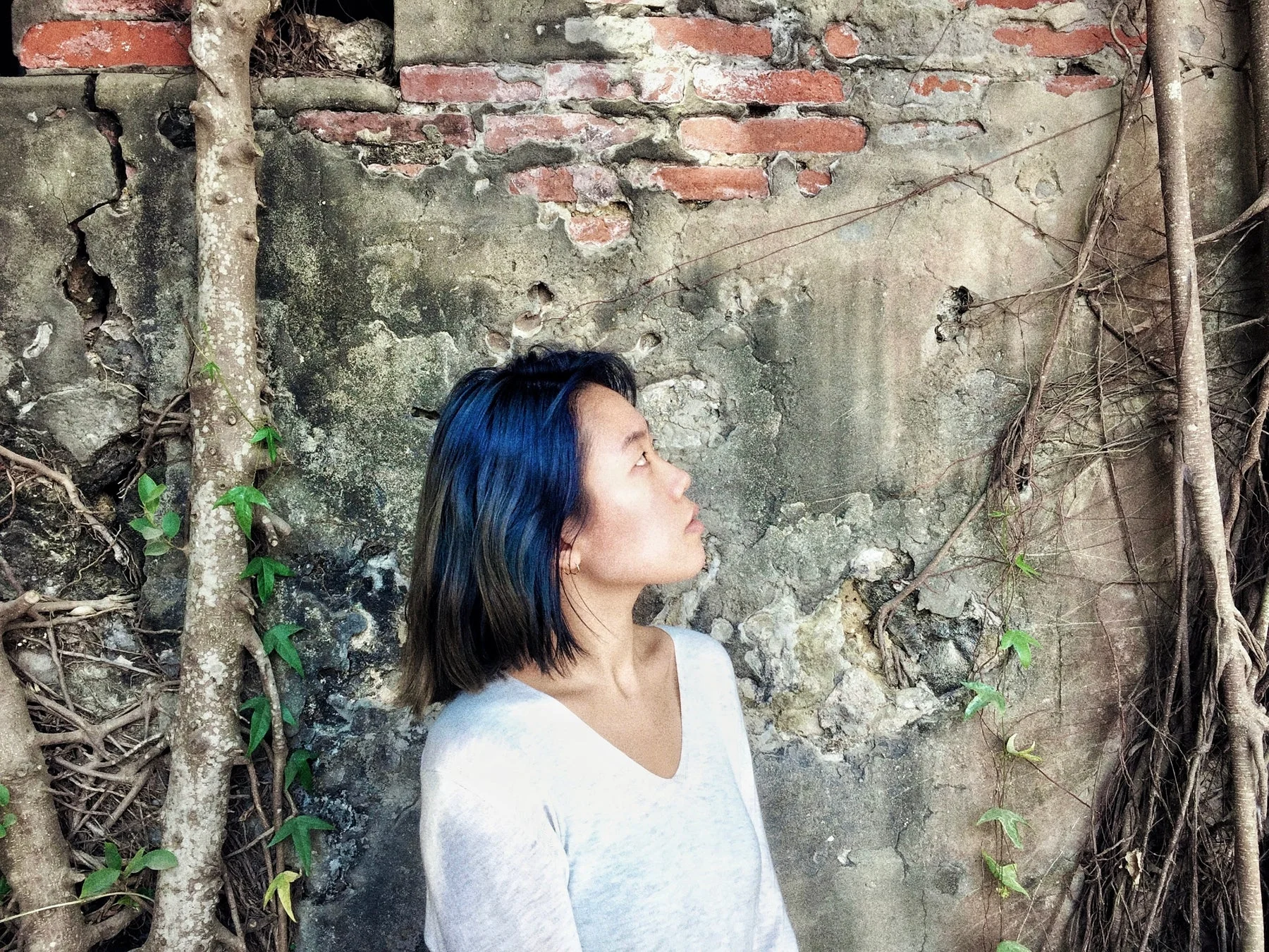 青い髪の女性が、木の根が絡まる古びた壁の前で上を見上げている横顔のポートレート。