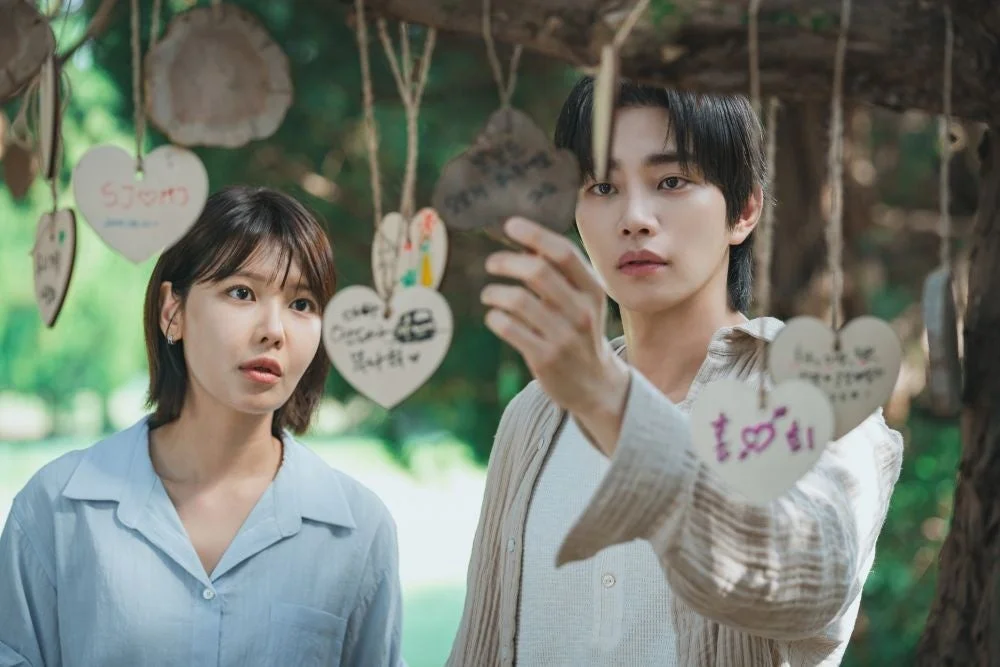 屋外で、男女が木に吊るされたハート形のプレートを見ているシーン。男性は手元のプレートに注目し、女性も隣でじっと見つめている。恋愛や願い事を連想させるロマンチックな雰囲気です。