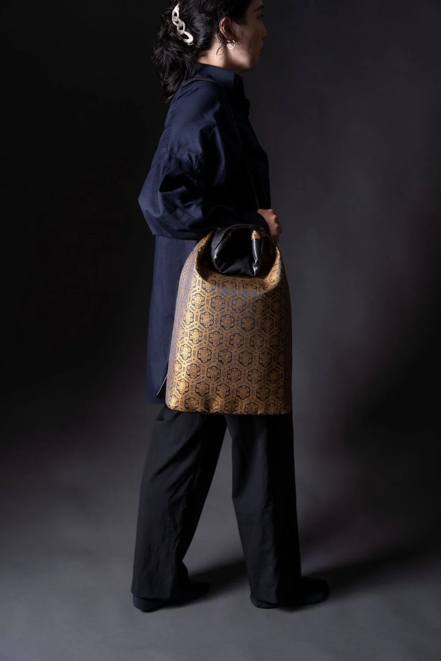 紺色のシャツと黒いパンツを着用した女性が、金色と黒の幾何学模様が特徴的なバッグを手にしている全身像