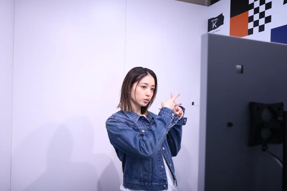 デニムジャケットを着た若い女性が、白い壁の前で指を交差させるポーズをしています。右側にはモニターの一部が見え、背景にはロゴと幾何学模様が配されています。
