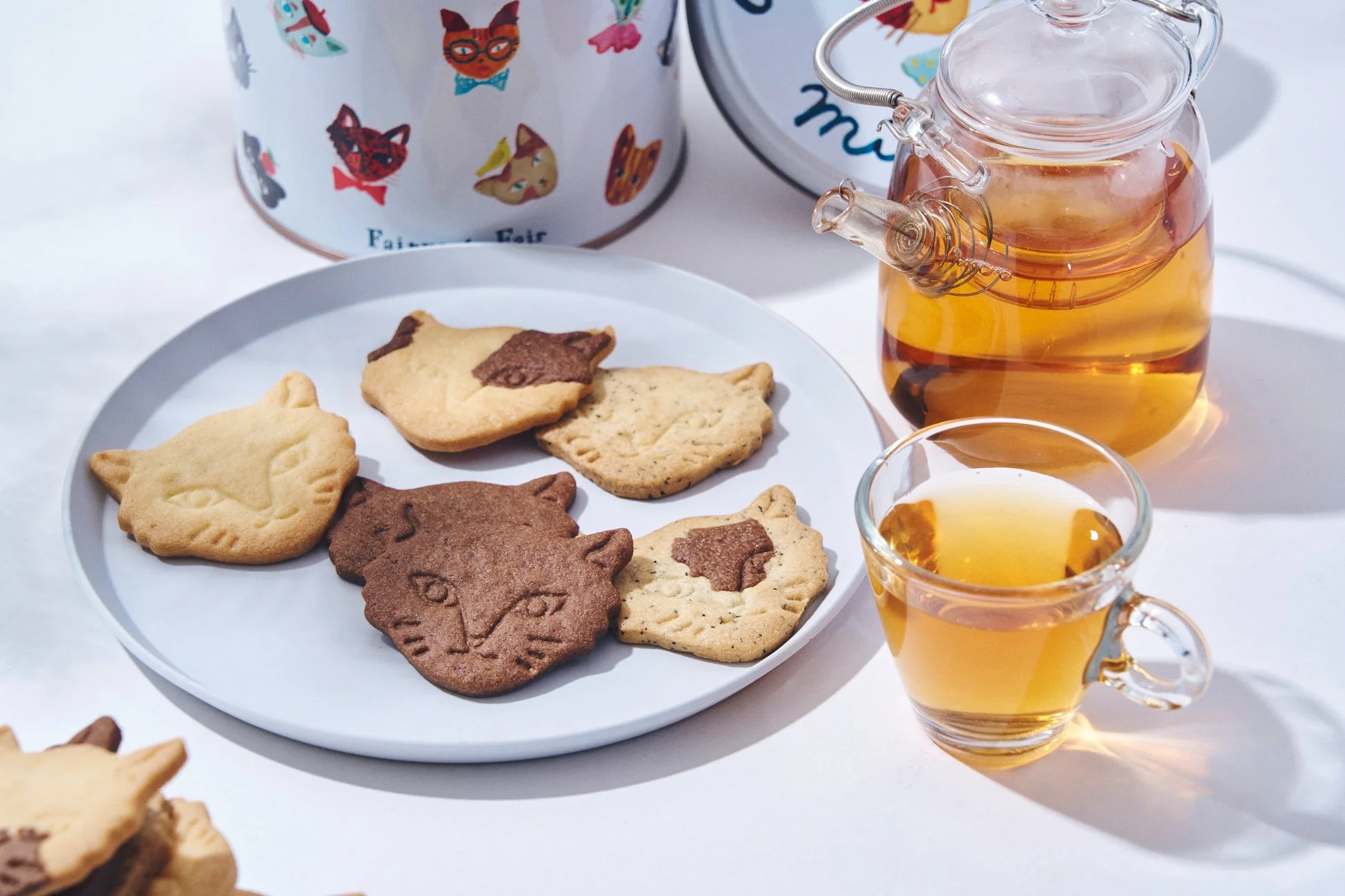 猫の形をした様々なフレーバーのクッキーが白いお皿に並べられ、ガラスのティーポットとティーカップ