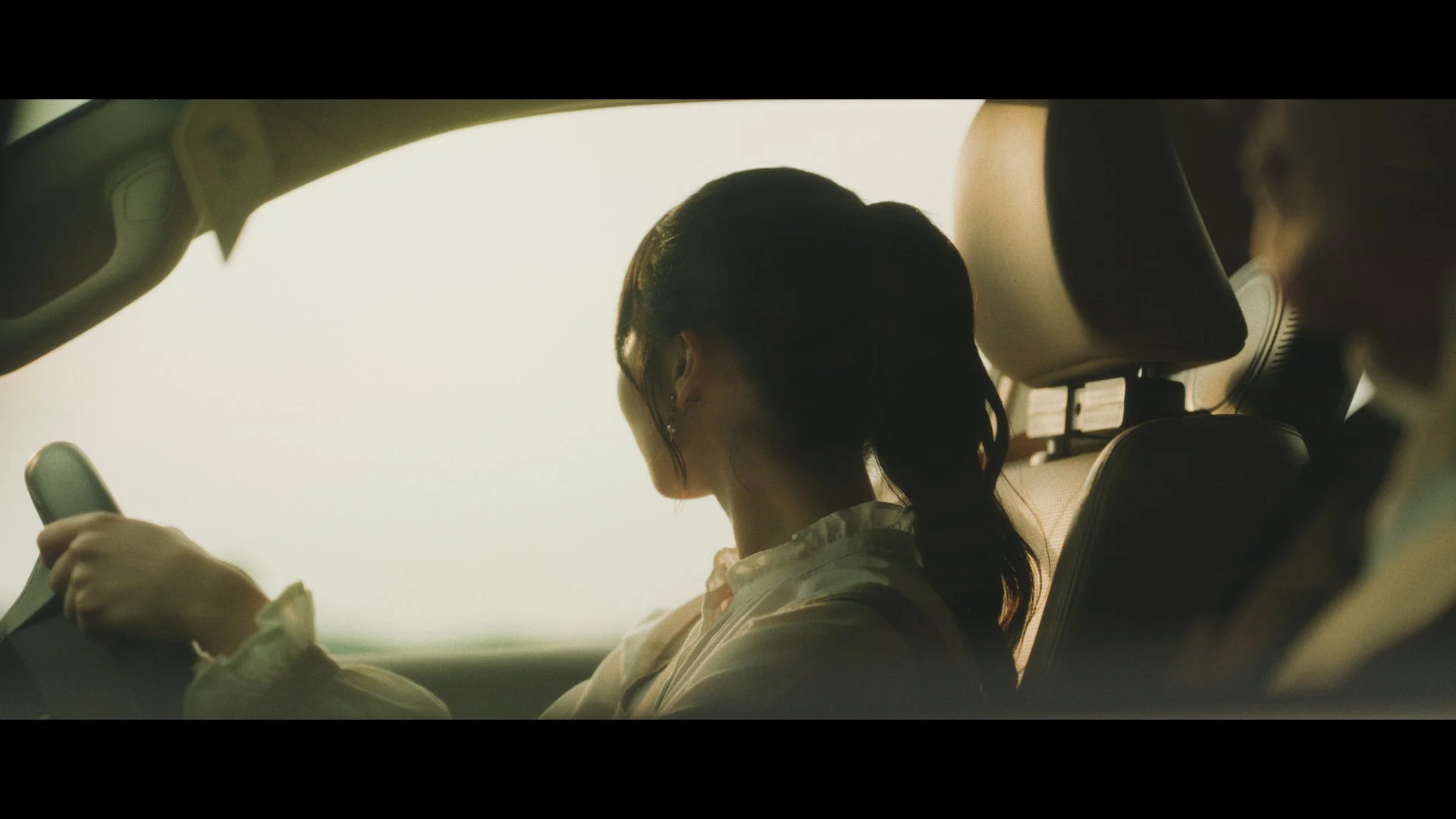 車を運転する女性の後ろ姿が写っています。夕暮れ時のような暖かい光が車内を照らし、運転席に座る女性の髪はポニーテールにまとめられています。静かで落ち着いた雰囲気です。