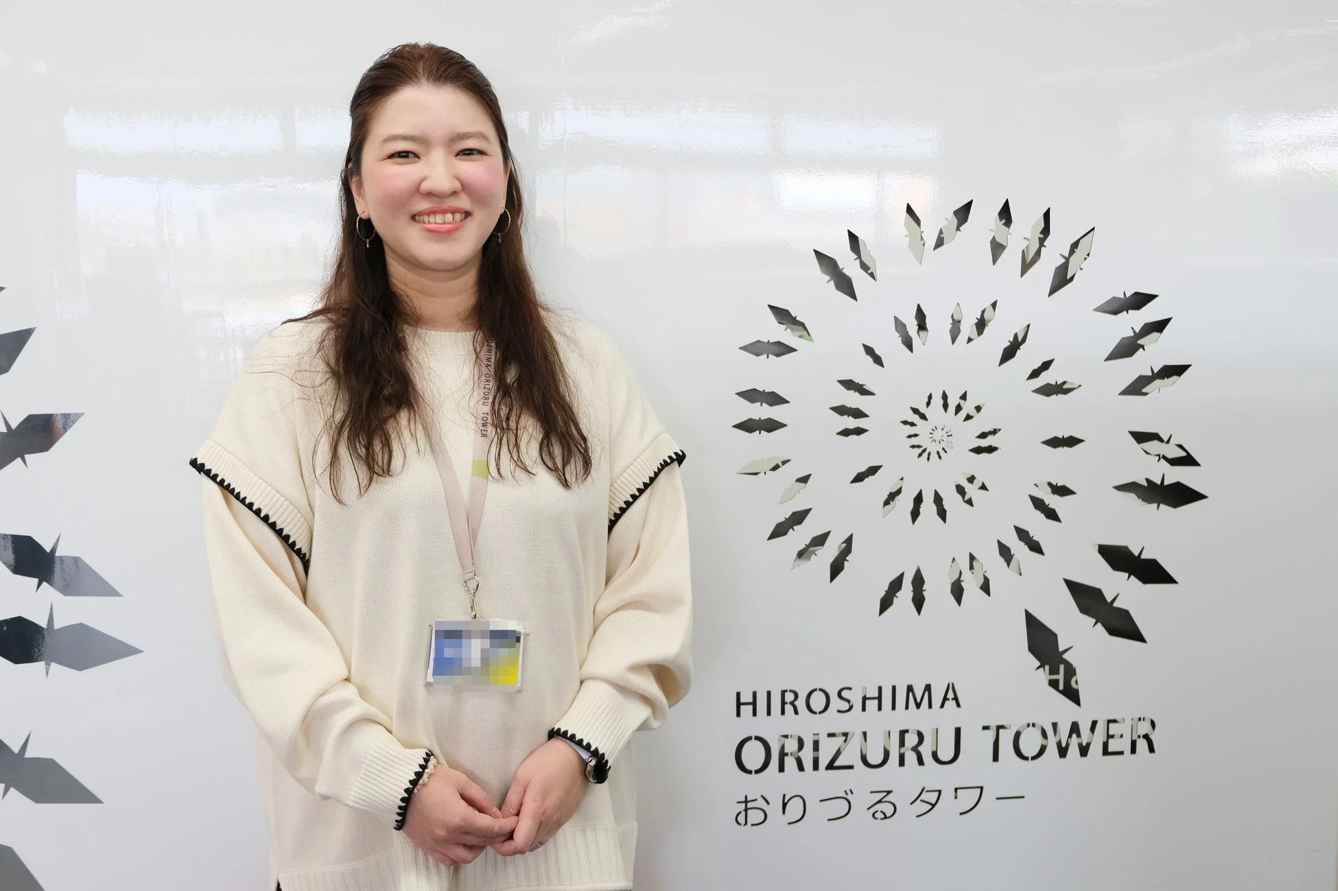 広島おりづるタワーのロゴと折り鶴の模様が描かれた白い壁の前で笑顔で立つ女性